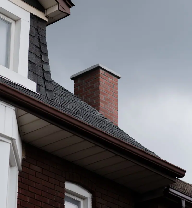 Brick chimney with a flat metal cap above an asphalt shingle roof and gutter