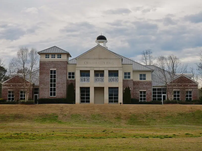 A brick and cream civic building with a small domed cupola and "CHELSEA CITY HALL" over the entrance, set behind a wide winter-brown lawn under a cloudy sky.