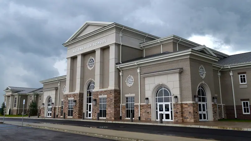The stone-and-brick front of a school with "HELENA HIGH SCHOOL" lettered on the pediment, arched windows and a wet forecourt under heavy grey storm clouds.