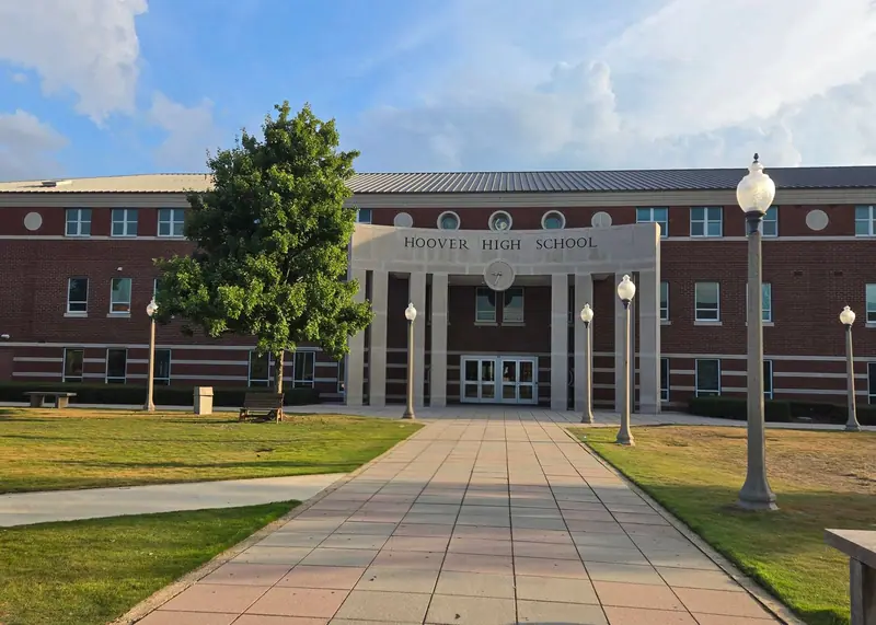 The brick and stone front entrance of a school with "HOOVER HIGH SCHOOL" lettered over the portico, a paved walk, lamp posts, lawn and a green tree in late-day sun.
