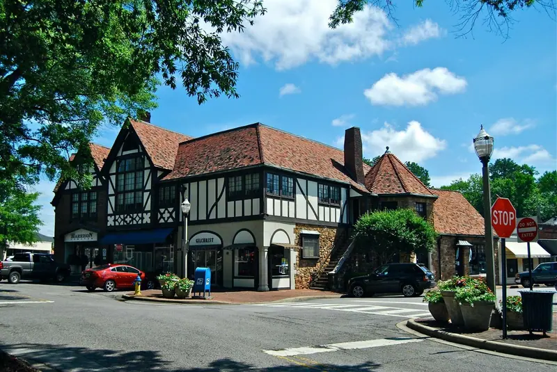 A two-storey Tudor-style half-timbered commercial building with a red tile roof and stone base on a village street corner, with shopfronts signed "Harrison Limited" and "Gilchrist", parked cars and flower planters under a blue sky.