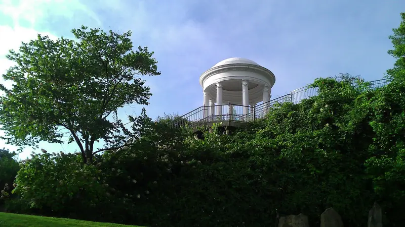 A white domed, columned round temple with an iron railing on a rise above thick green shrubs and a tree, against a hazy blue sky; phone-camera quality, and three dark upright stone slabs sit in the bottom-right corner.